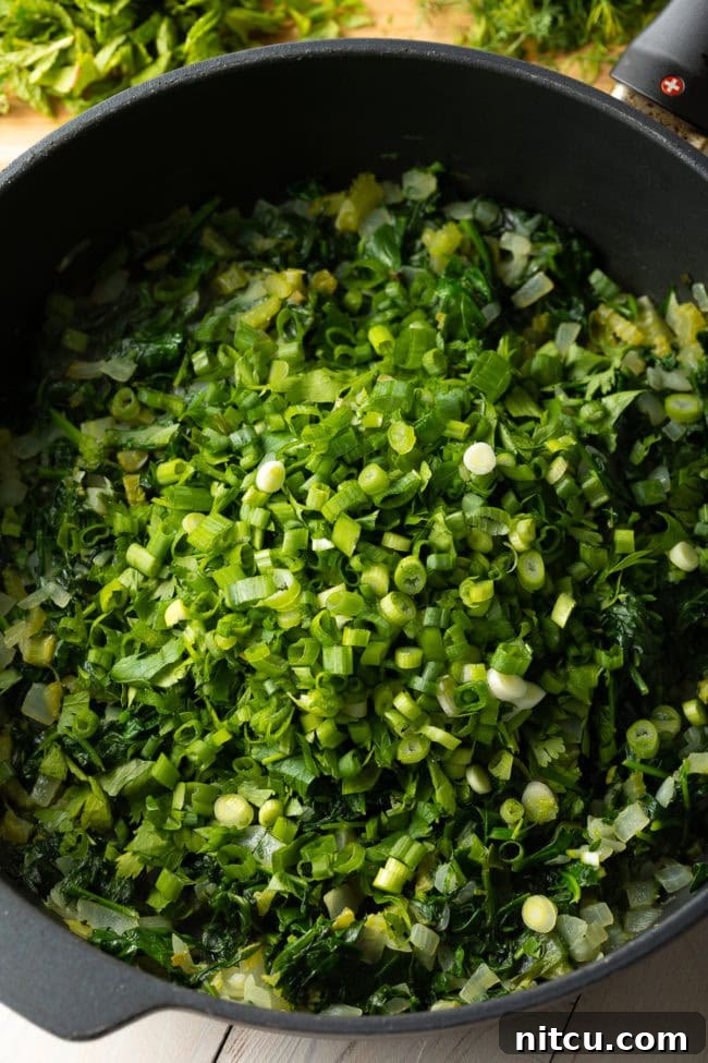 A large pot of vibrant green vegetables, including wilted spinach and freshly added scallions, continuing to cook down for the pie filling.