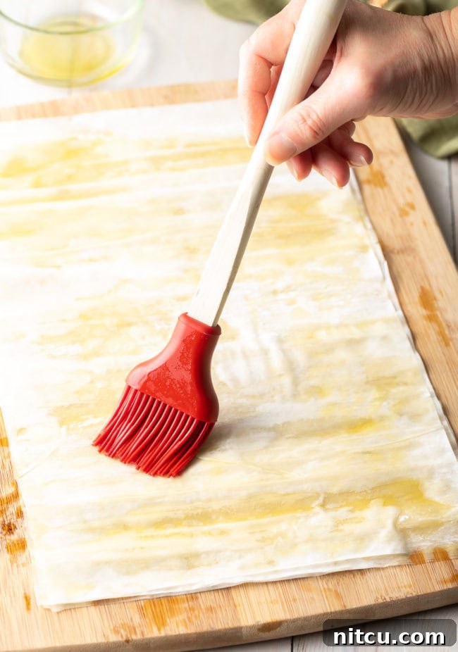 A hand expertly brushing a thin, even layer of melted ghee onto a single sheet of delicate phyllo dough, preparing it for layering.