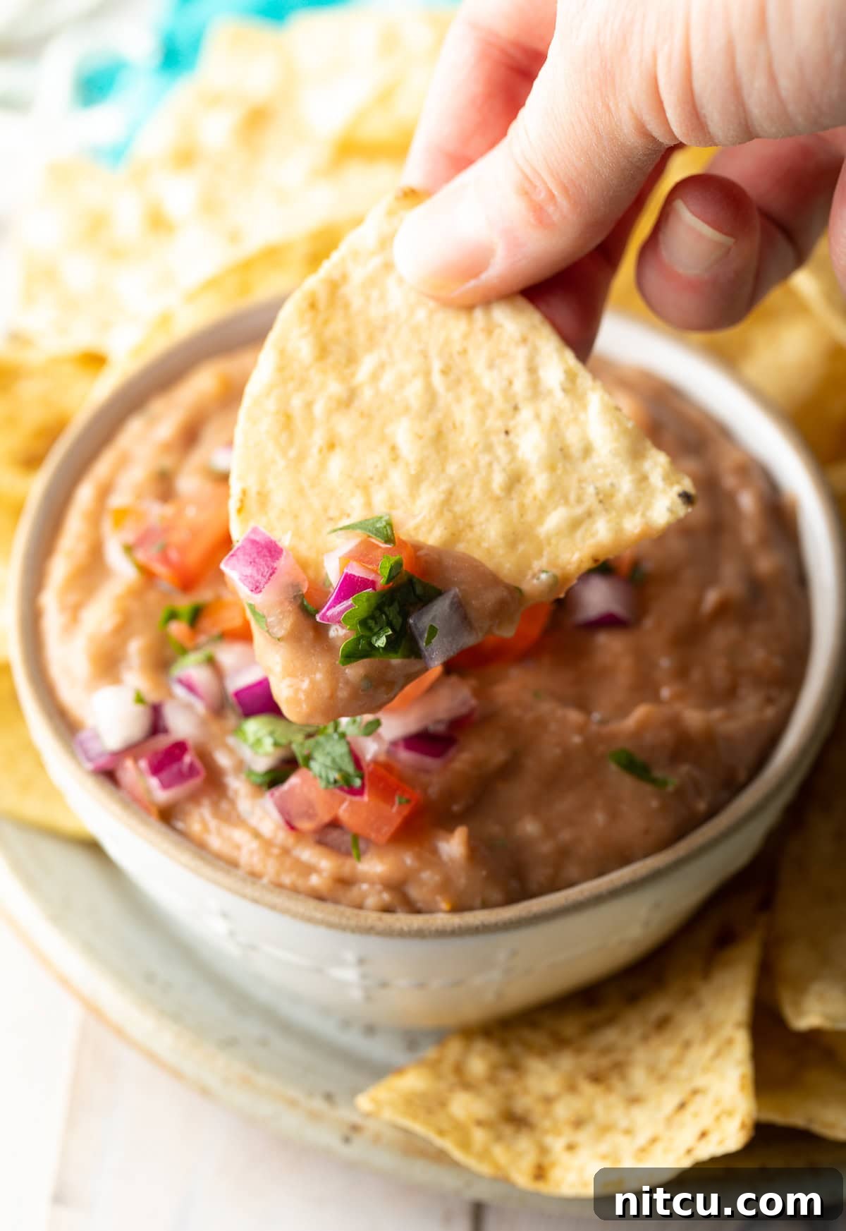 A hand holding a golden tortilla chip, poised to scoop a generous portion of creamy, flavorful homemade bean dip.