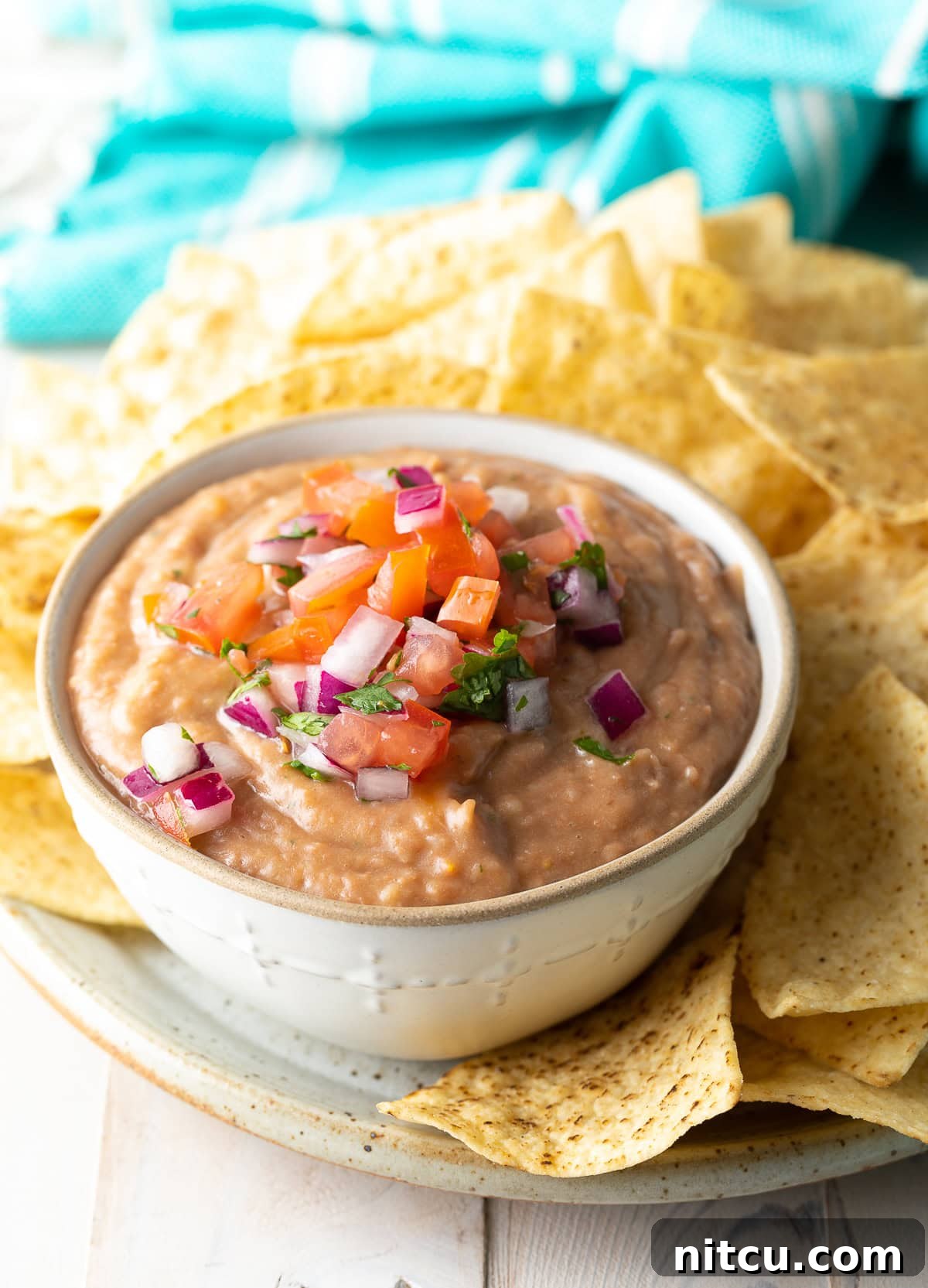 A pristine white bowl filled with creamy homemade bean dip, artfully garnished, positioned next to a rustic plate brimming with crunchy tortilla chips.
