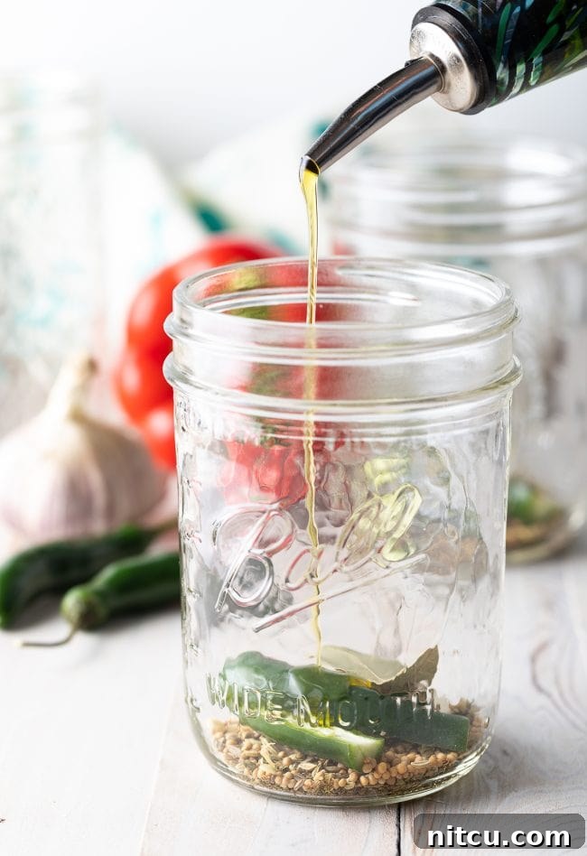 A hand pouring olive oil over a mix of spices and serrano chili in a jar, preparing for giardiniera.