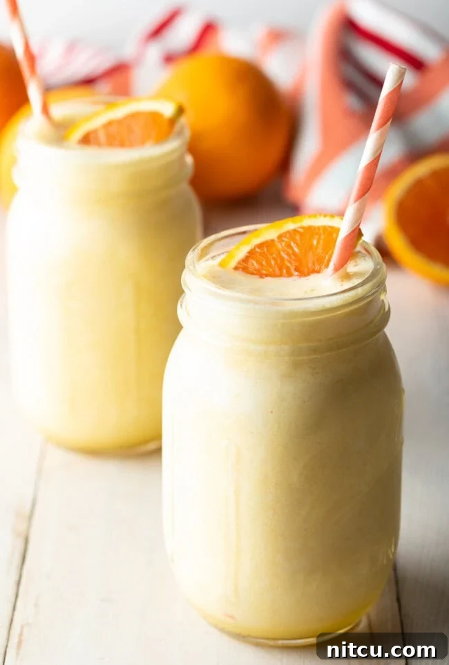 Two jars filled with frothy orange Julius smoothies, garnished with fresh orange slices and straws, sitting on a light-colored surface.