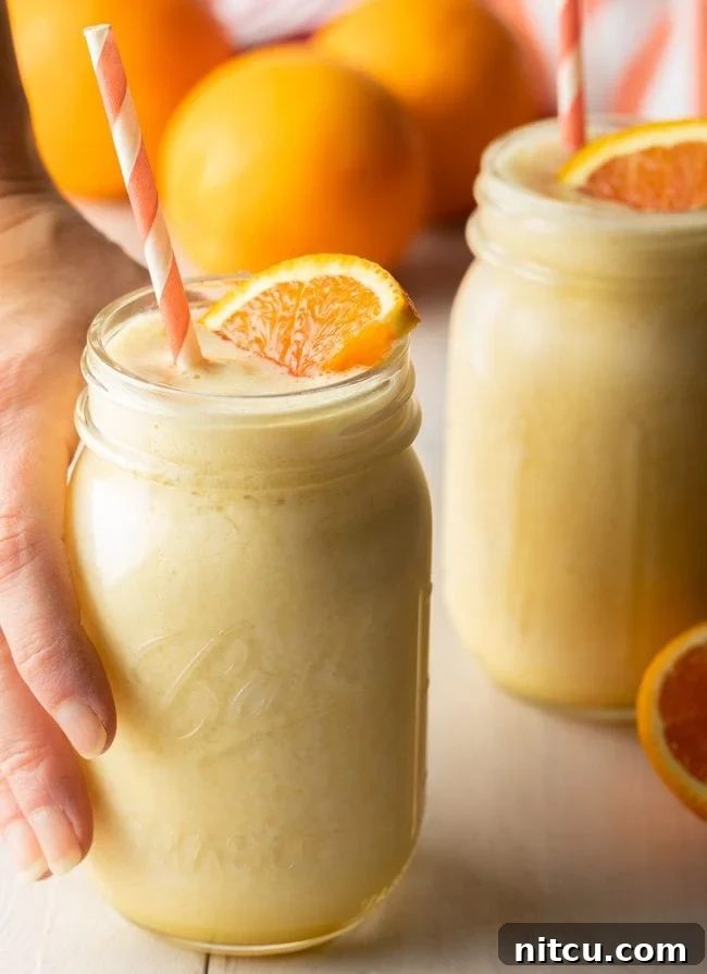 A hand reaching for a jar filled with a frothy Orange Julius smoothie, garnished with a fresh orange slice.