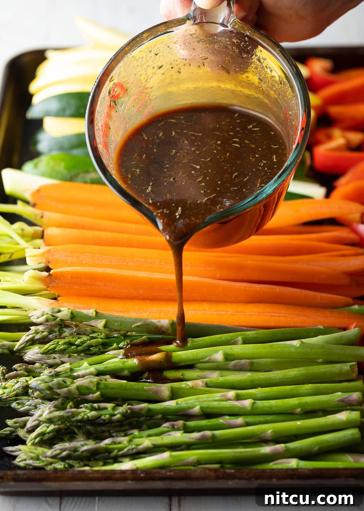 Pouring the homemade marinade over raw, cut vegetables on a baking sheet, ready for grilling.
