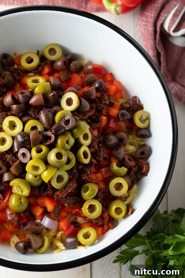 A bowl filled with uncooked orzo, kalamata olives, sun-dried tomatoes, and diced red bell peppers, ready to be mixed.