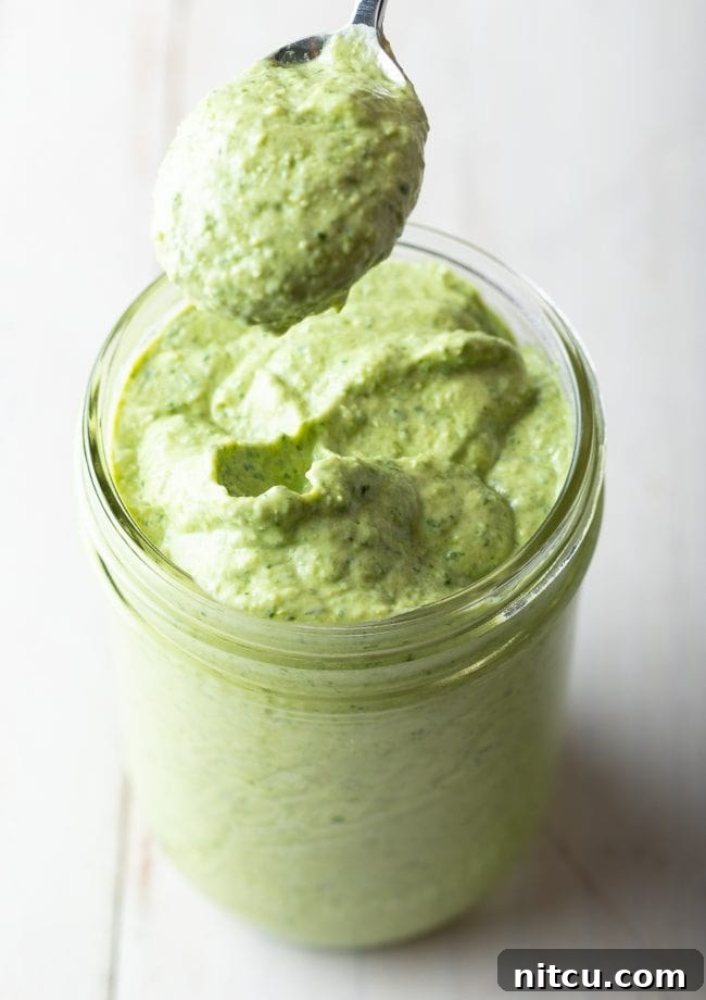 A clear glass jar brimming with smooth, green pesto cream sauce, with a spoon gently scooping a portion from its luscious surface.