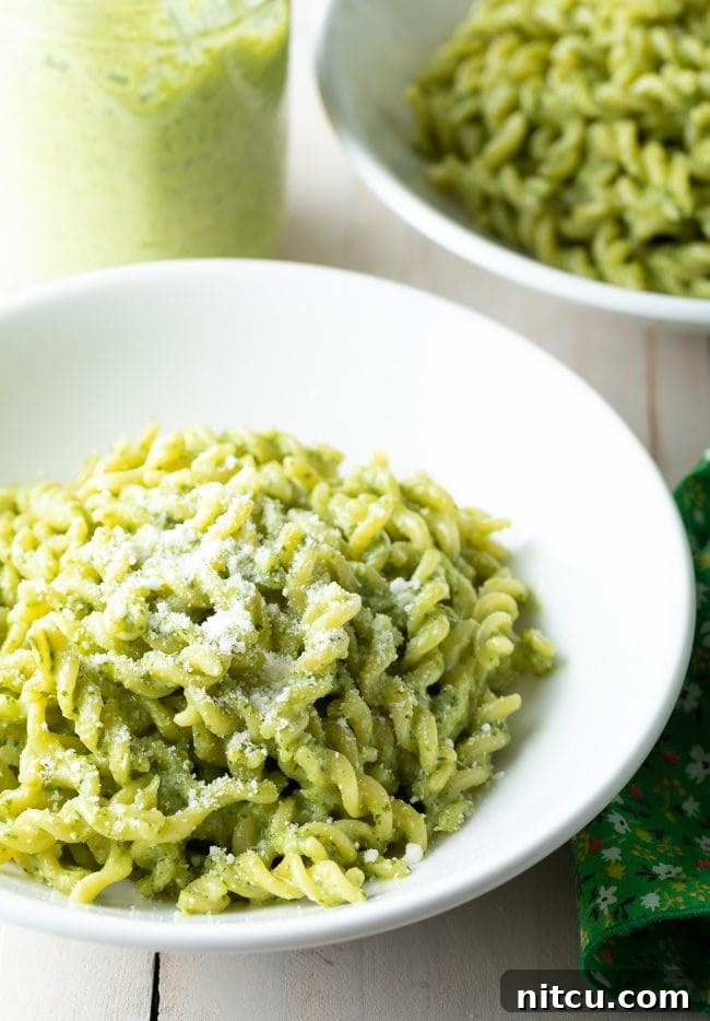 Two white bowls filled with creamy pesto pasta, alongside a jar of the fresh pesto sauce, arranged on a light surface.