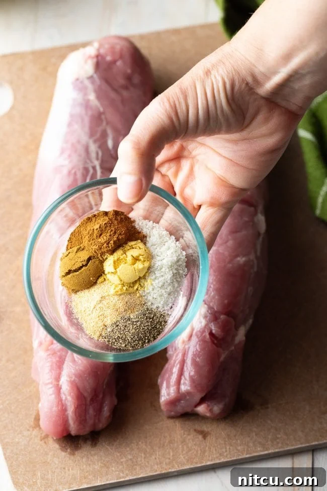 Two raw pork tenderloins on a cutting board with a hand holding a small bowl with spices above them.