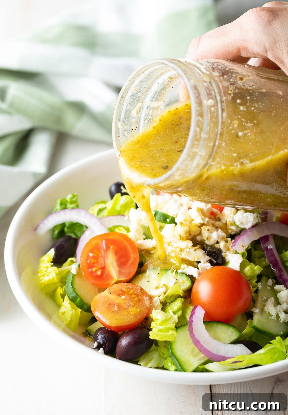A hand pouring a glass of Greek Salad Dressing over a Greek Salad, highlighting its fresh appeal.