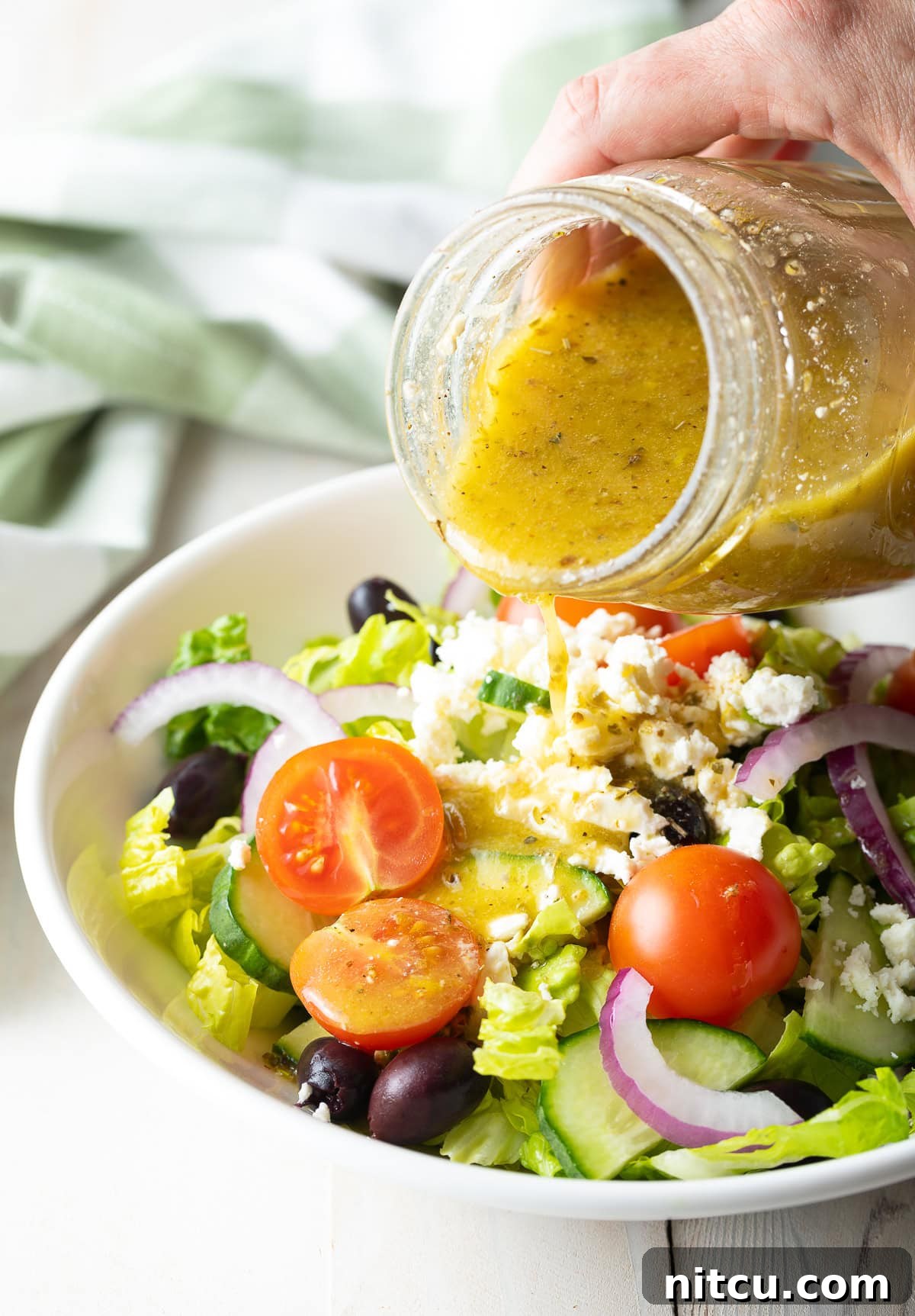 A hand pouring a glass of Greek Salad Dressing over a Greek Salad, ready for serving.