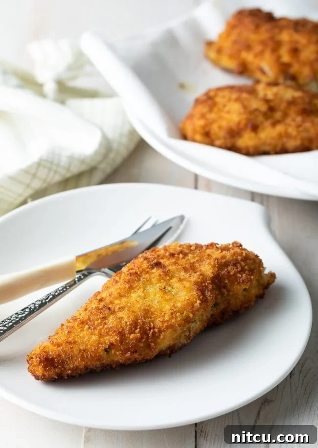 A perfectly golden-brown, crispy pan-fried Chicken Kiev served elegantly on a pristine white plate, accompanied by a fork, highlighting its exquisite texture and readiness to be savored.
