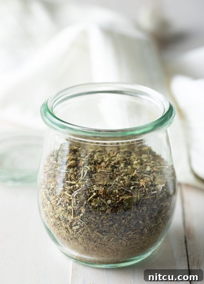 A hand holding a spoon filled with homemade Italian seasoning over a jar of the blend.