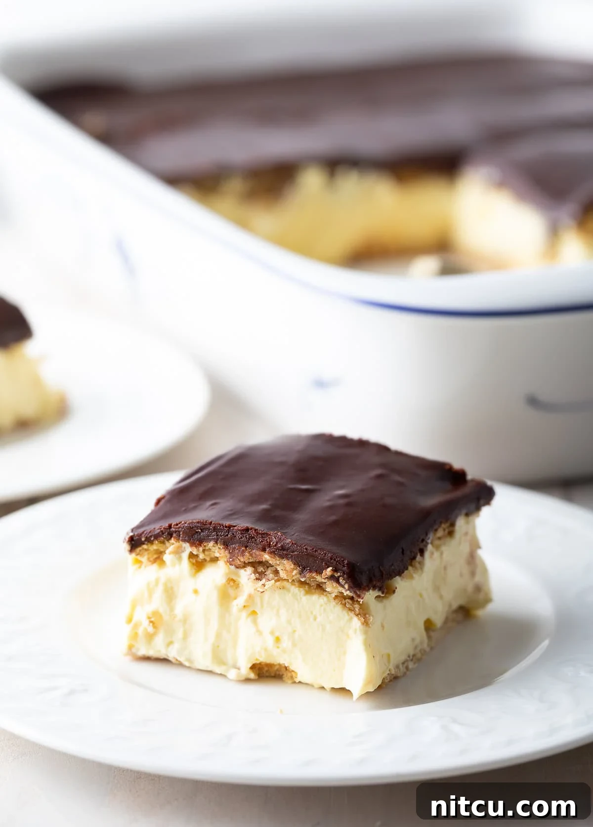 A perfectly portioned slice of no-bake eclair cake, showcasing its distinct layers of softened graham cracker, creamy vanilla pudding, and rich chocolate ganache, artfully presented on a white plate, ready to be enjoyed.