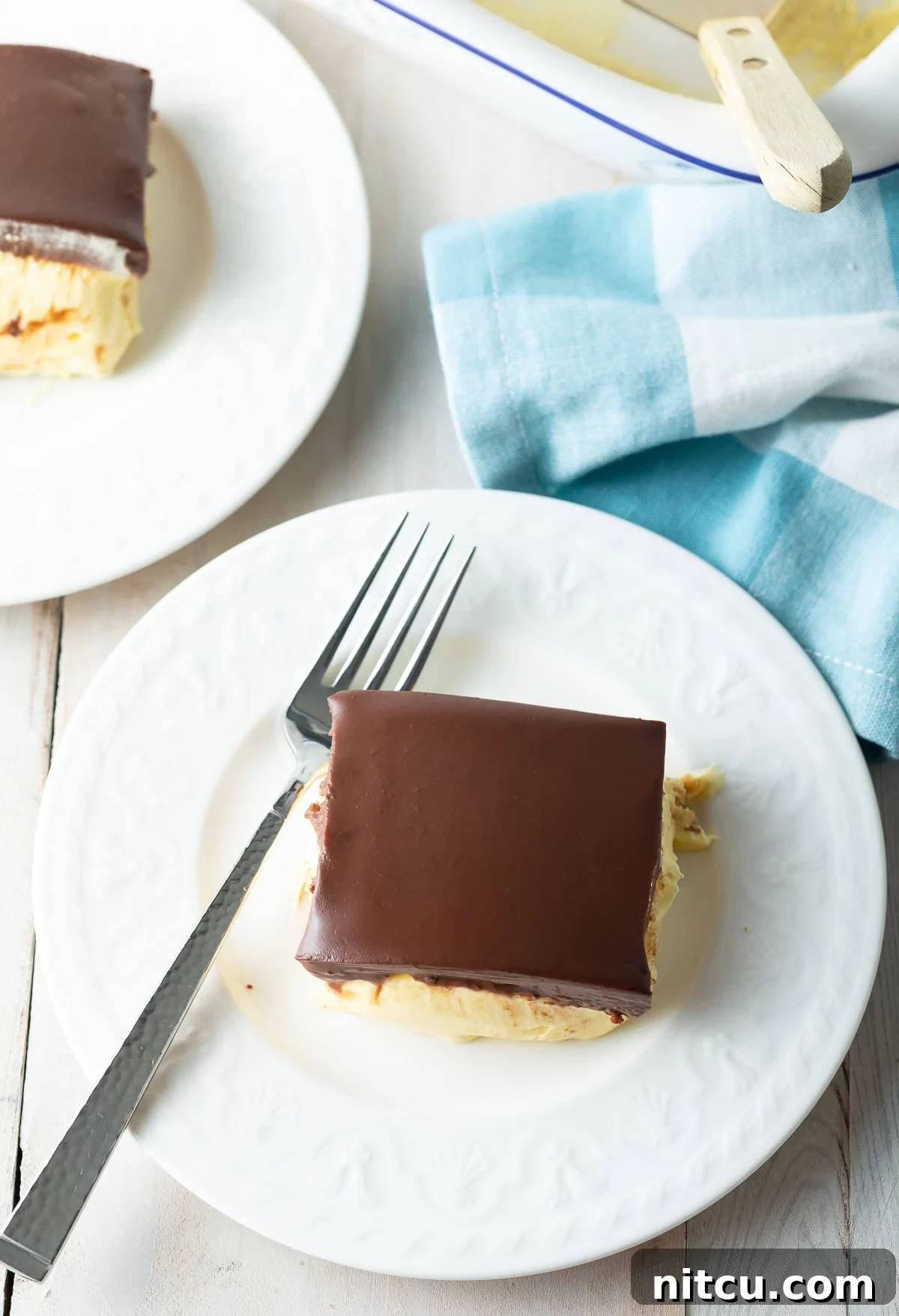 An overhead shot of an entire no-bake eclair cake with chocolate ganache, sliced and ready to serve on plates with elegant blue napkins, set on a rustic wooden table, highlighting its rich, inviting presentation.