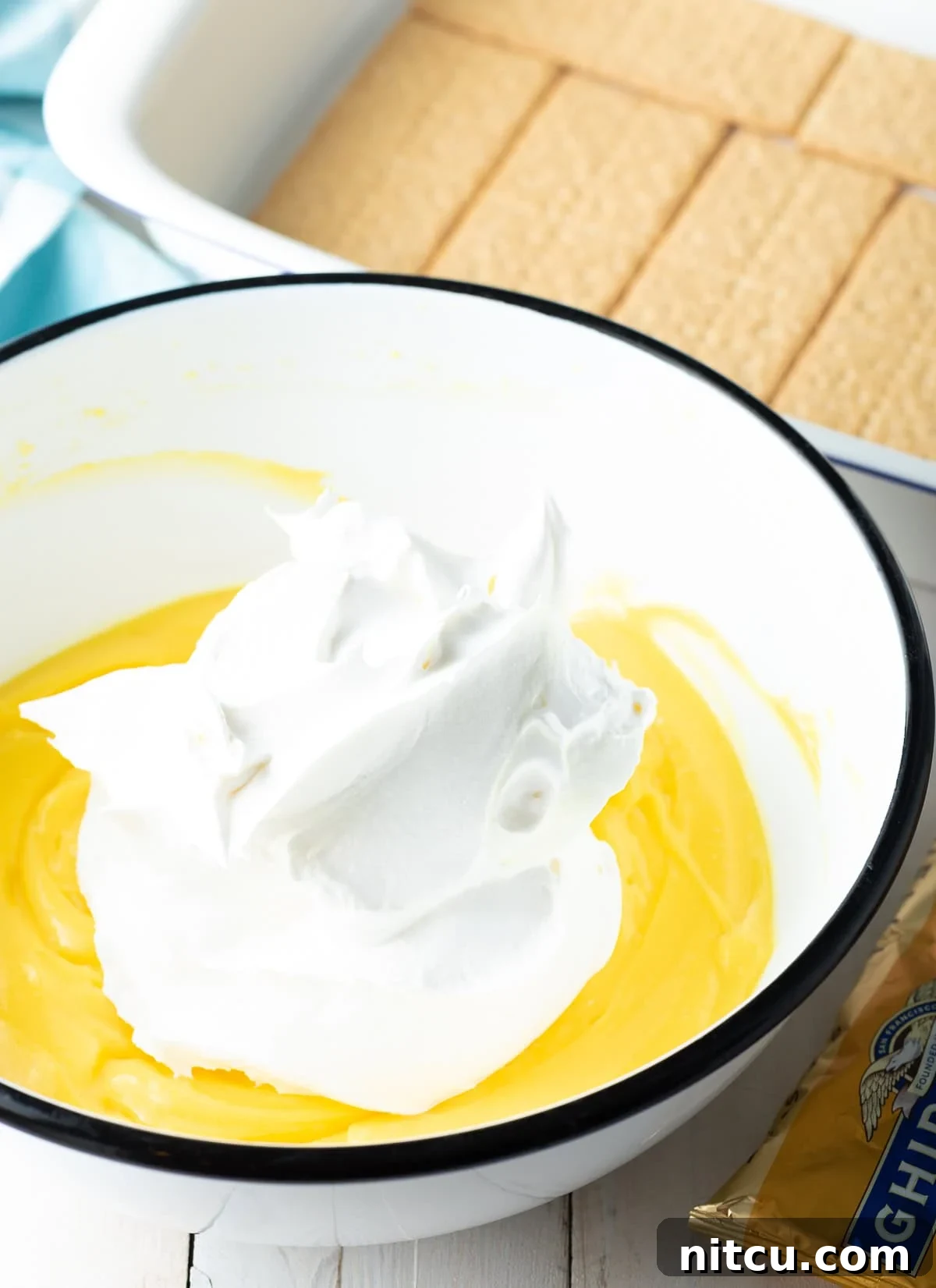 A mixing bowl filled with creamy vanilla pudding, with a generous dollop of fluffy Cool Whip carefully placed on top, ready to be folded in and create the luscious eclair cake filling.