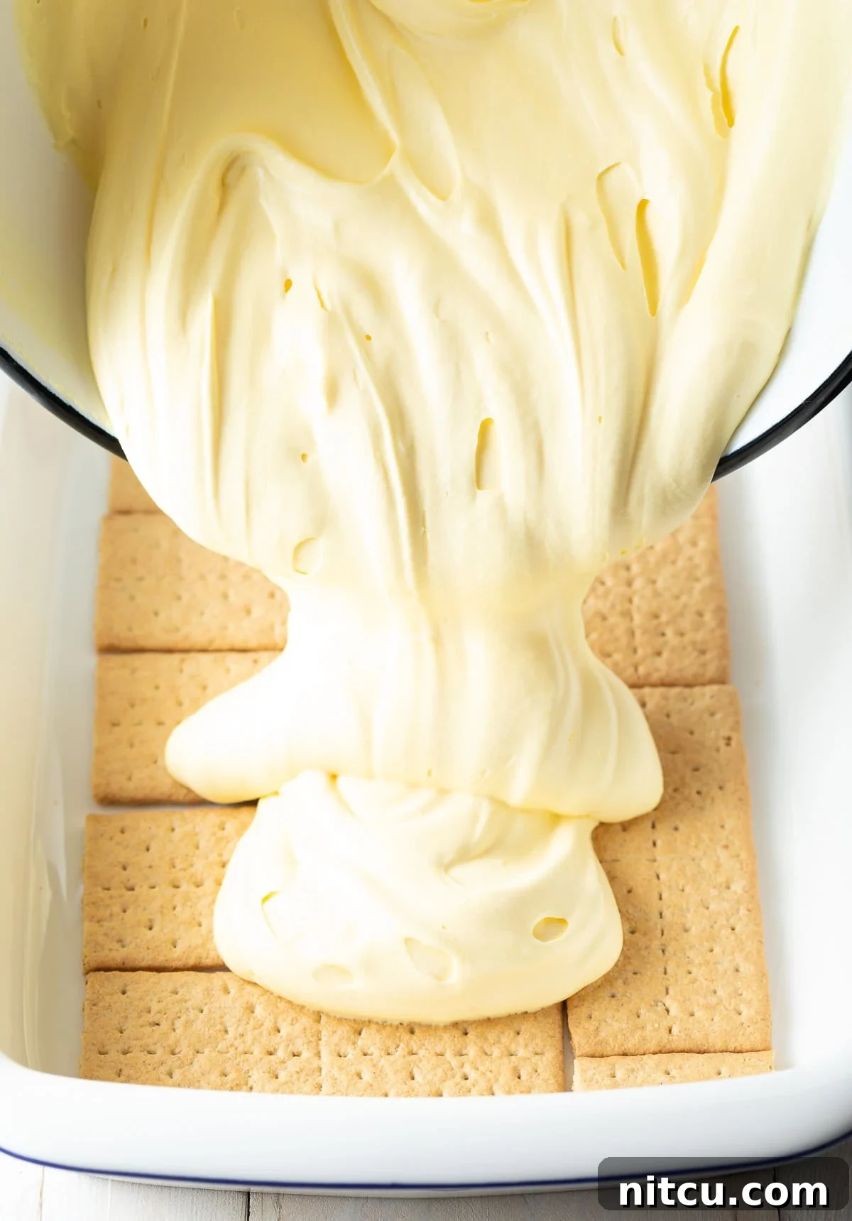 A baking tray meticulously lined with a single layer of graham crackers, with the smooth, creamy pudding mixture being poured evenly over them, forming the luxurious second layer of the no-bake eclair cake.