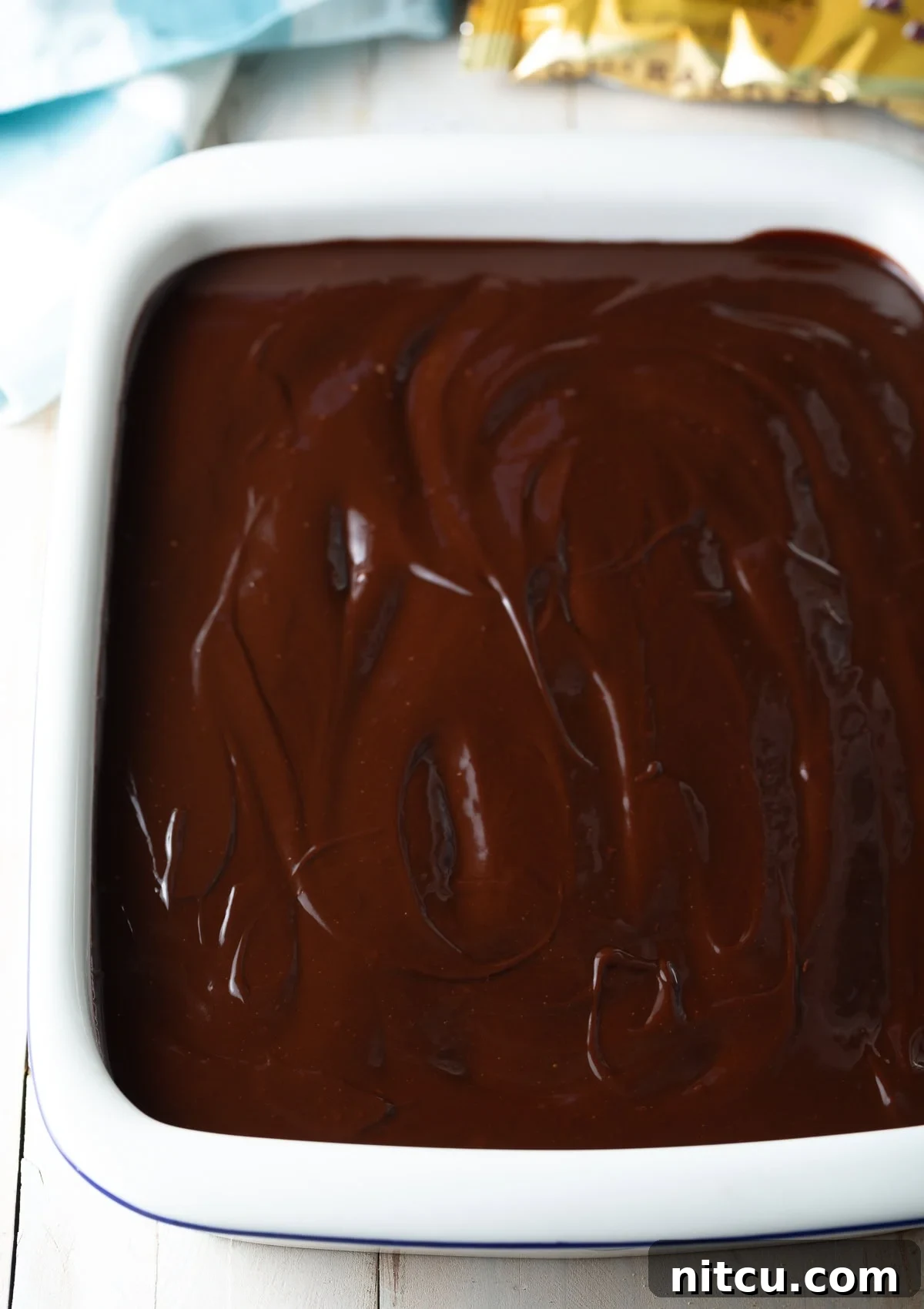 A top-down view of the no-bake eclair cake nestled in a baking dish, freshly topped with a rich, glossy chocolate ganache, which is being smoothly spread to cover the entire surface, promising a decadent finish.