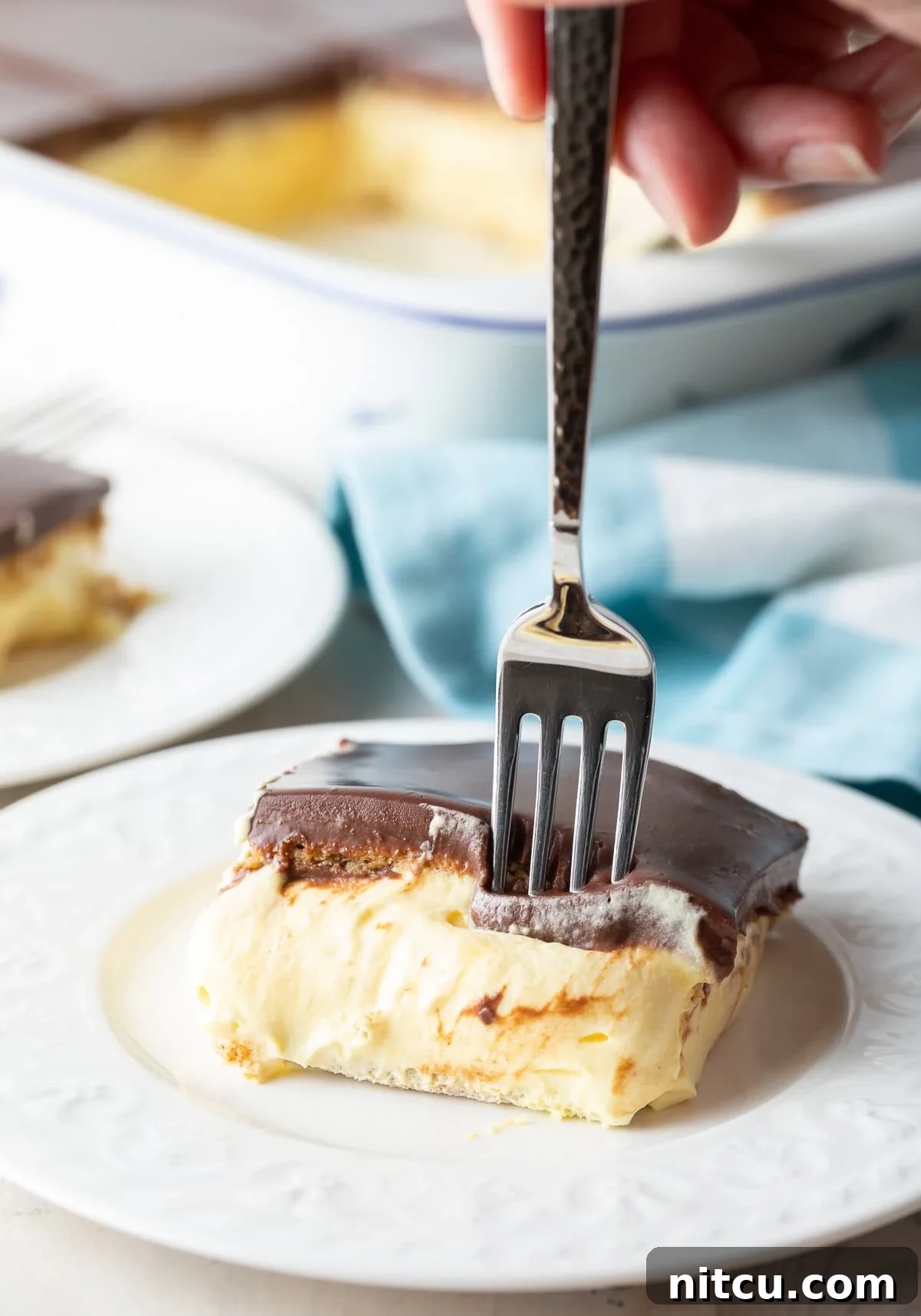 A close-up side view of a fork gently entering a slice of the no-bake eclair cake, highlighting the beautiful layers of chocolate ganache, creamy filling, and softened graham cracker, tempting a bite.