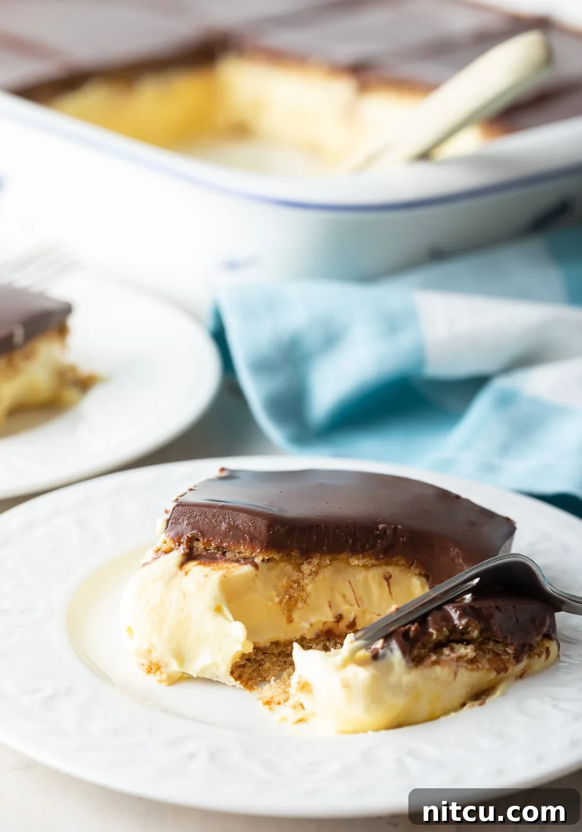 A fork lifts a generous slice of the layered no-bake eclair cake from a plate, showcasing the creamy filling and thick chocolate ganache, inviting diners to indulge in its rich texture.