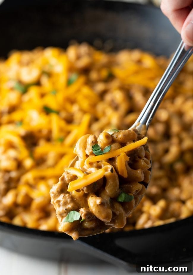 A metal spoon lifts a generous serving of creamy cheeseburger macaroni from a skillet, highlighting the rich cheese and tender pasta.