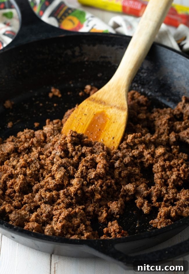 Ground beef browning in a large skillet, being broken apart with a wooden spatula.