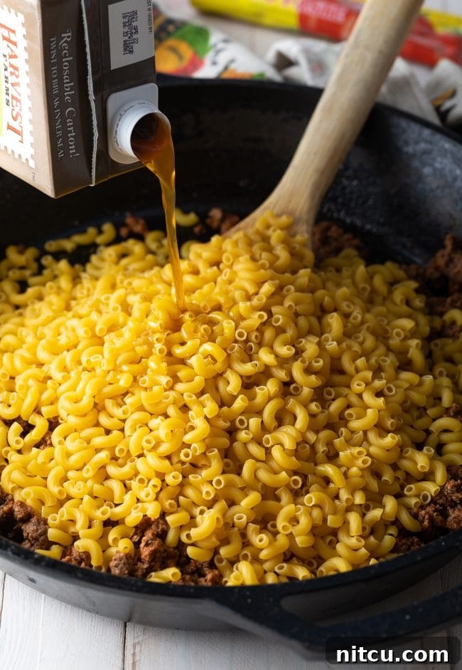 Beef broth being poured from a carton into a skillet with uncooked macaroni noodles and seasoned ground beef.