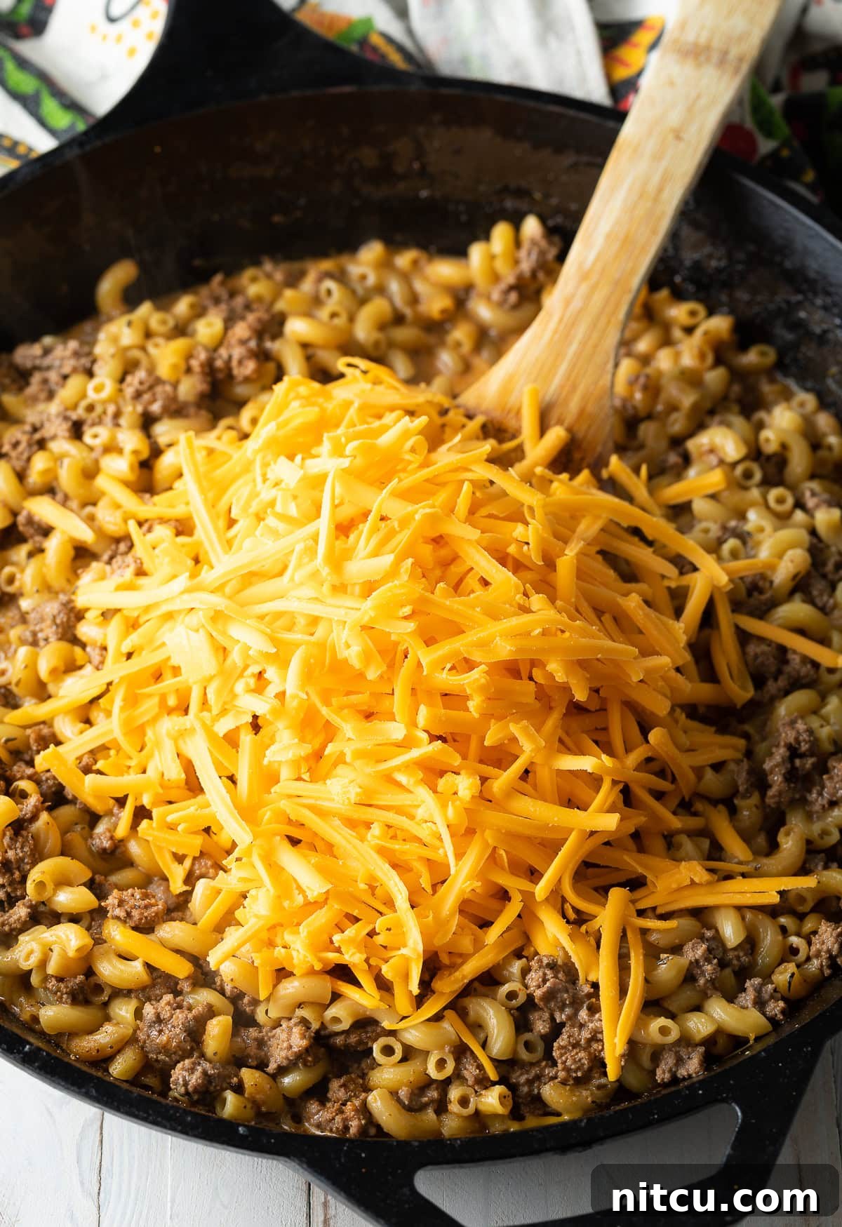 A hand stirring shredded cheddar cheese into the simmering beef and macaroni skillet, creating a creamy sauce.