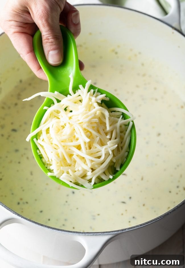 Hand holding a green measuring cup filled with shredded mozzarella cheese, ready to be added to the sauce.