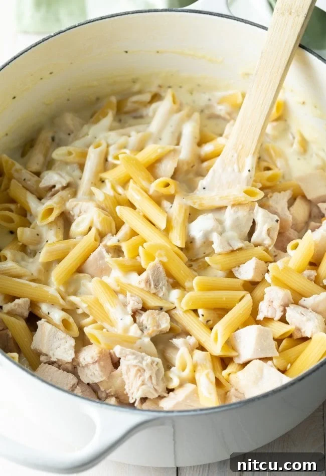 Large cooking pot showing penne pasta, cooked chicken chunks, and a creamy Alfredo sauce being stirred with a wooden spoon.