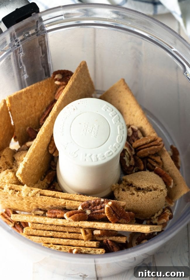 Graham crackers, pecans, and brown sugar measured out in a food processor, ready to be processed for the crust.