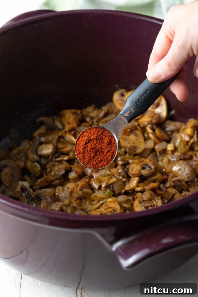 A close-up of smoked paprika being added to sautéed mushrooms and onions in a pot.