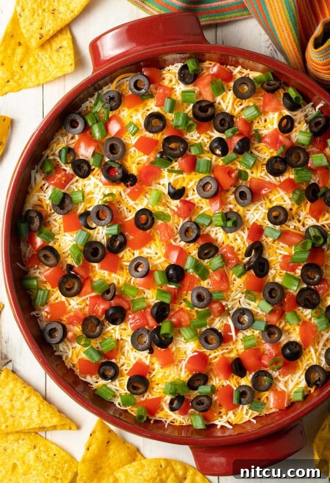 Top down view red round dish with layered Mexican taco pizza, shredded cheese, chopped green onions, and sliced black olives, and chopped tomatoes. Surrounded by tortilla chips.