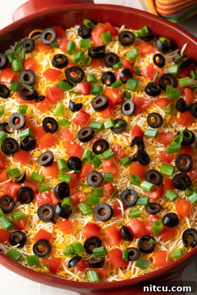 Top down view layered taco pizza dip with shredded cheddar cheese, sliced green onions, sliced black olives, and chopped tomatoes.