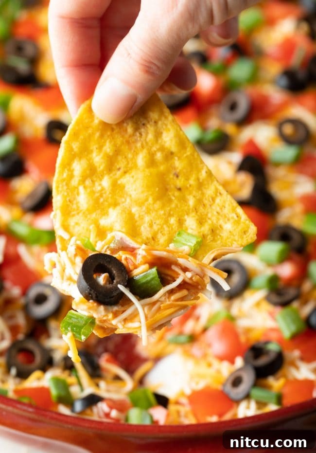 Hand using a tortilla chip to scoop into Mexican pizza dip with shredded cheddar cheese, sliced green onions, sliced black olives, and chopped tomatoes.