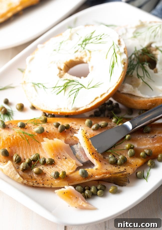 A close-up shot of flaky smoked trout being gently pulled apart with a knife on a plate, next to a bagel.
