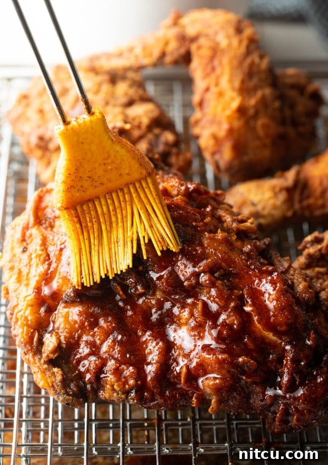 A brush applying vibrant red spicy oil evenly over the surface of a fried chicken piece.