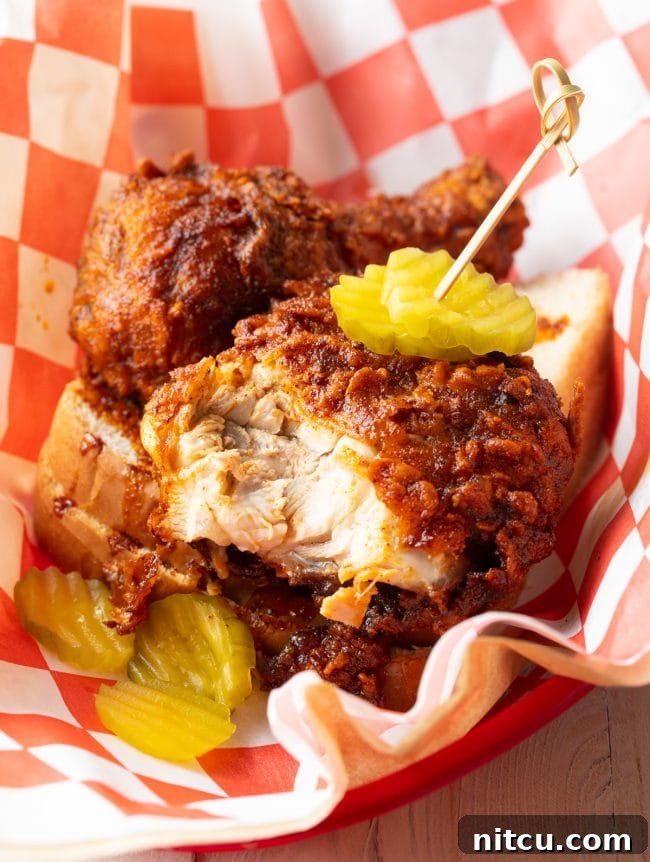 A piece of Nashville hot chicken, sliced open to reveal its tender, juicy breast meat, resting in a serving basket alongside fresh dill pickles.
