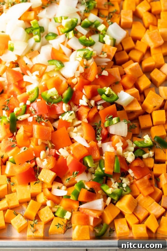 A large baking sheet filled with an array of vibrant, freshly chopped vegetables including sweet potatoes, bell peppers, onions, and jalapeños, ready for roasting.