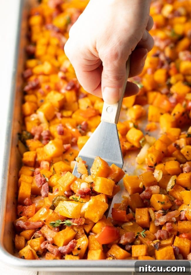 A spatula gently flipping the mixture of sweet potatoes and other vegetables on a baking sheet, ensuring even cooking and browning.