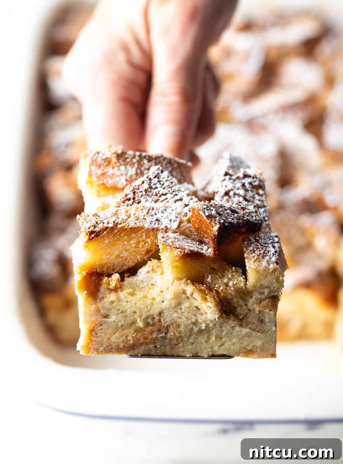 A slice of French toast casserole overnight recipe being held up with a spatula over a white baking dish.