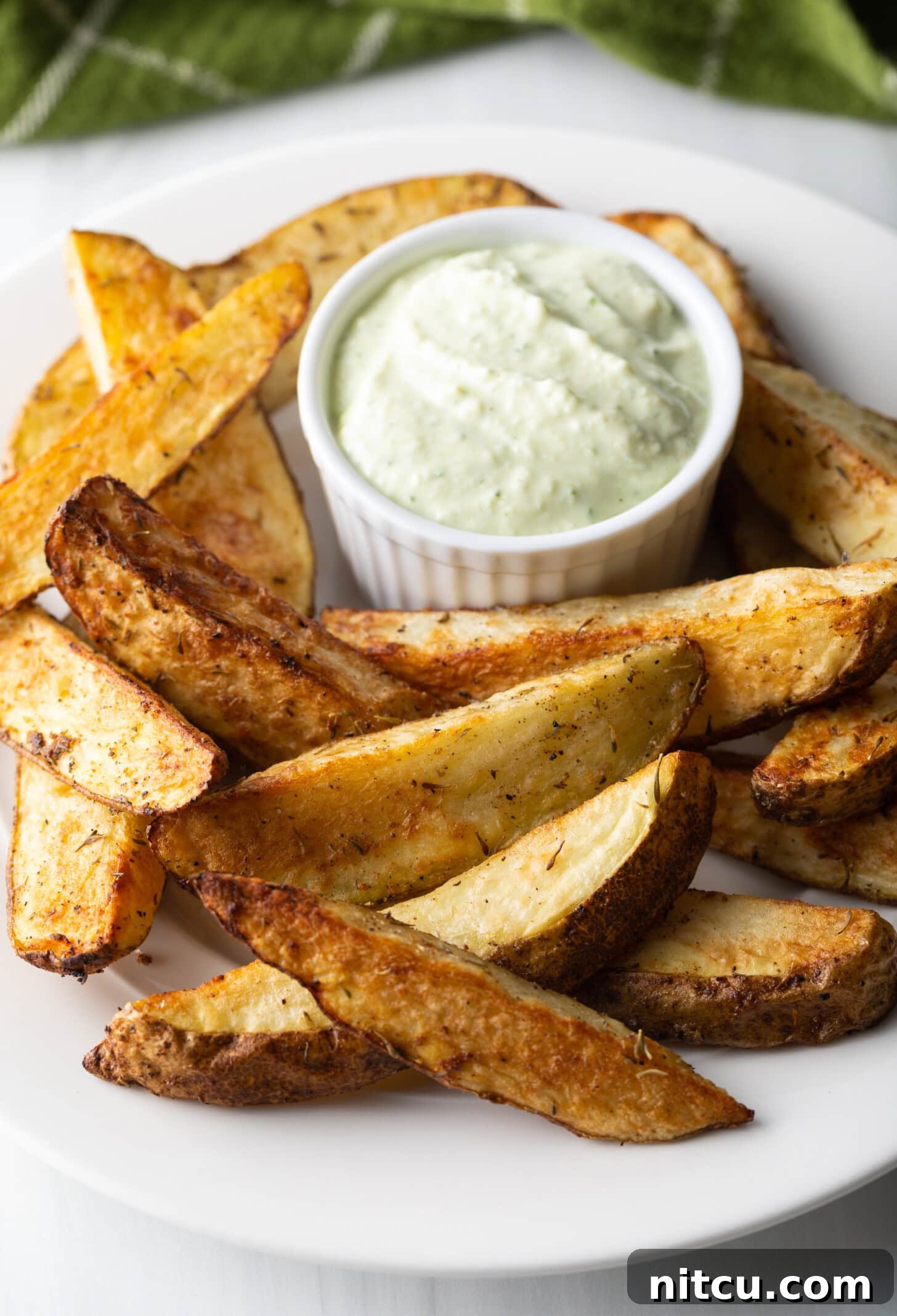 A white plate piled high with golden brown, thick-cut air fryer potato wedges, accompanied by a small white ramekin filled with a creamy white dipping sauce.
