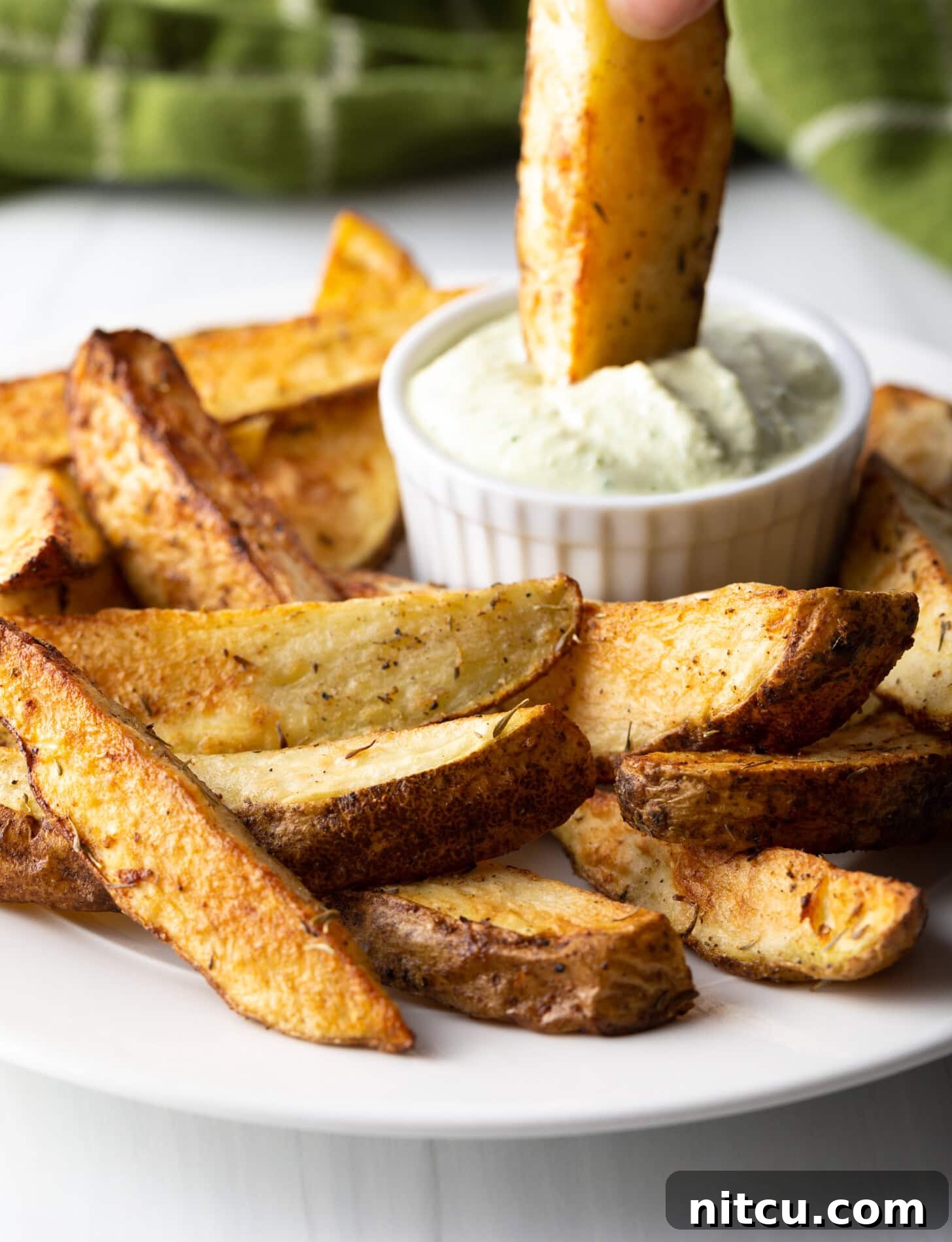 A hand gently dips a golden-brown, crispy air fryer potato wedge into a small white ramekin filled with creamy white dipping sauce.