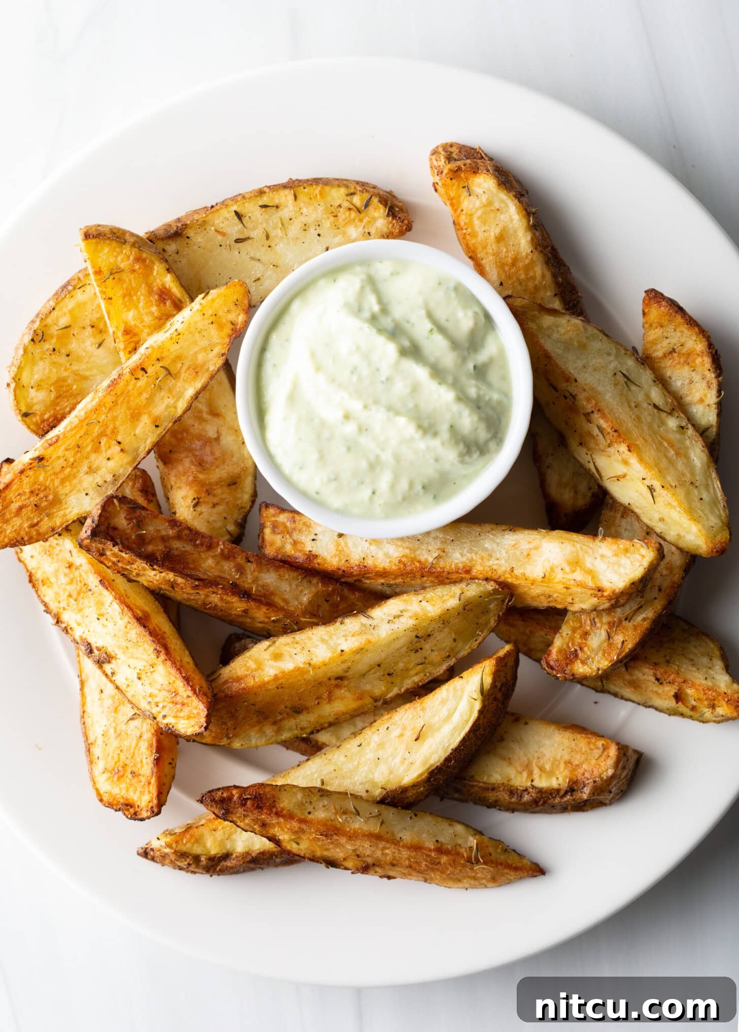 A white plate is generously stacked with thick-cut, golden-brown air fryer potato wedges, accompanied by a small white ramekin holding a creamy dipping sauce.