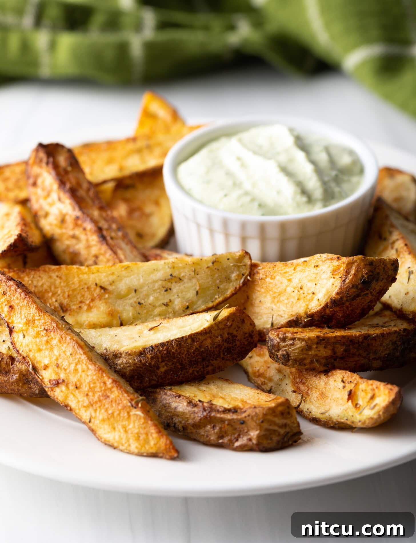 A beautiful side view of a white plate generously filled with thick-cut potato wedges, perfectly cooked in an air fryer, with a small white ramekin of dipping sauce alongside.