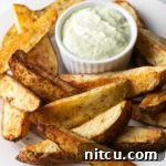 A white plate piled with thick-cut air fryer potato wedges, with a white ramekin of white dipping sauce also on the plate.