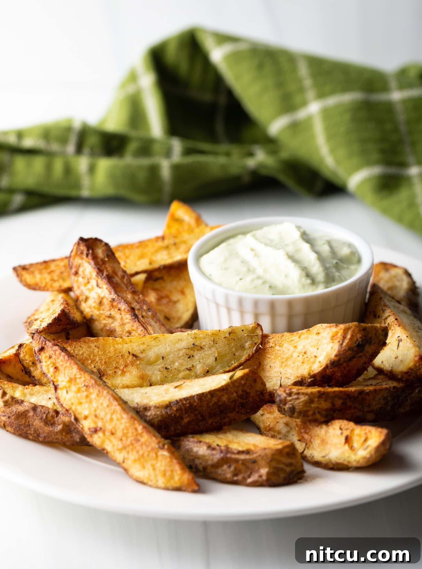 A pristine white plate showcases a generous serving of crispy potato wedges, freshly made in the air fryer. Beside them, a small white ramekin holds a creamy, light-colored dipping sauce.