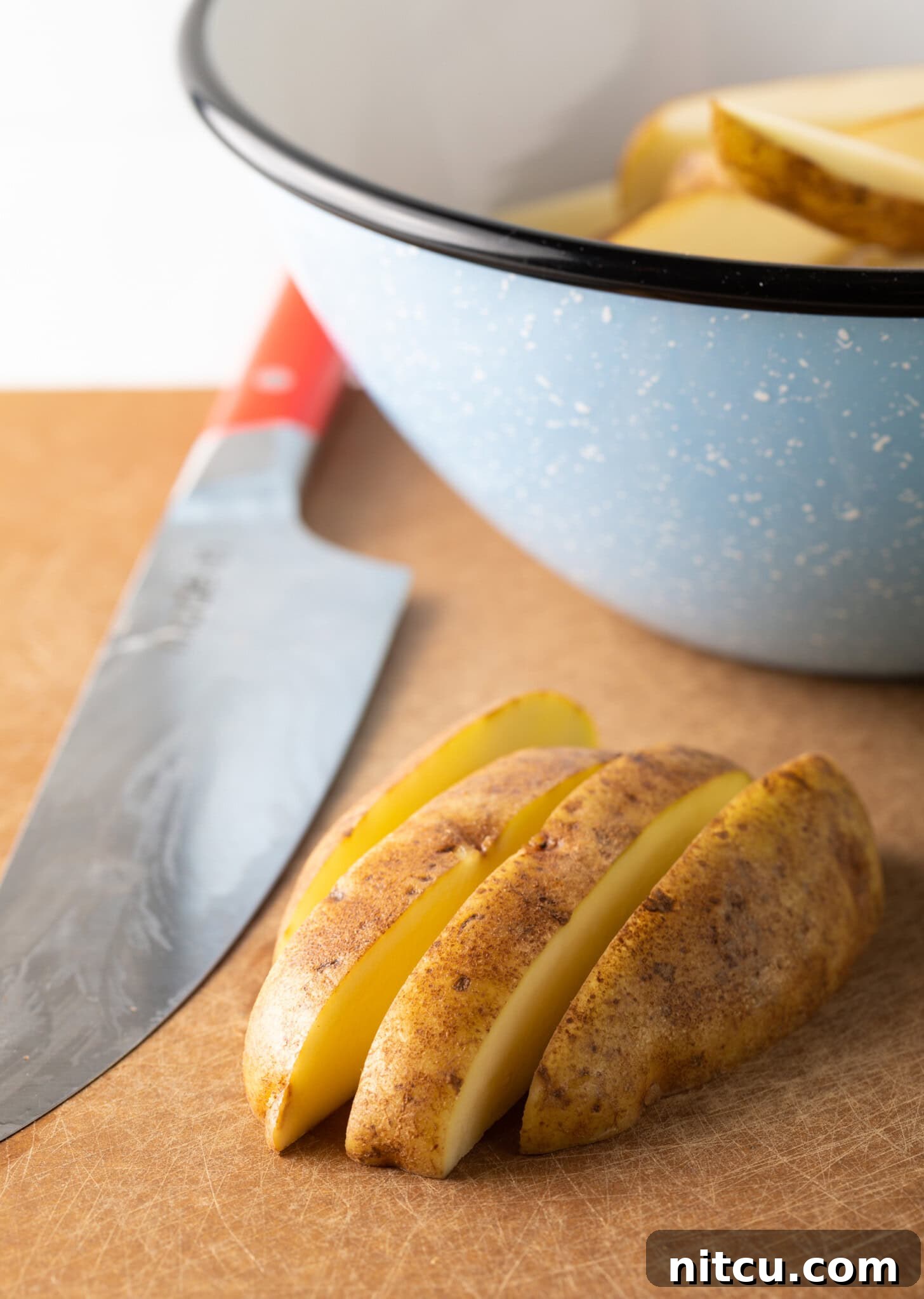 A cutting board holds a half-cut raw potato, skillfully sliced into thick, uniform wedges with a sharp knife nearby.