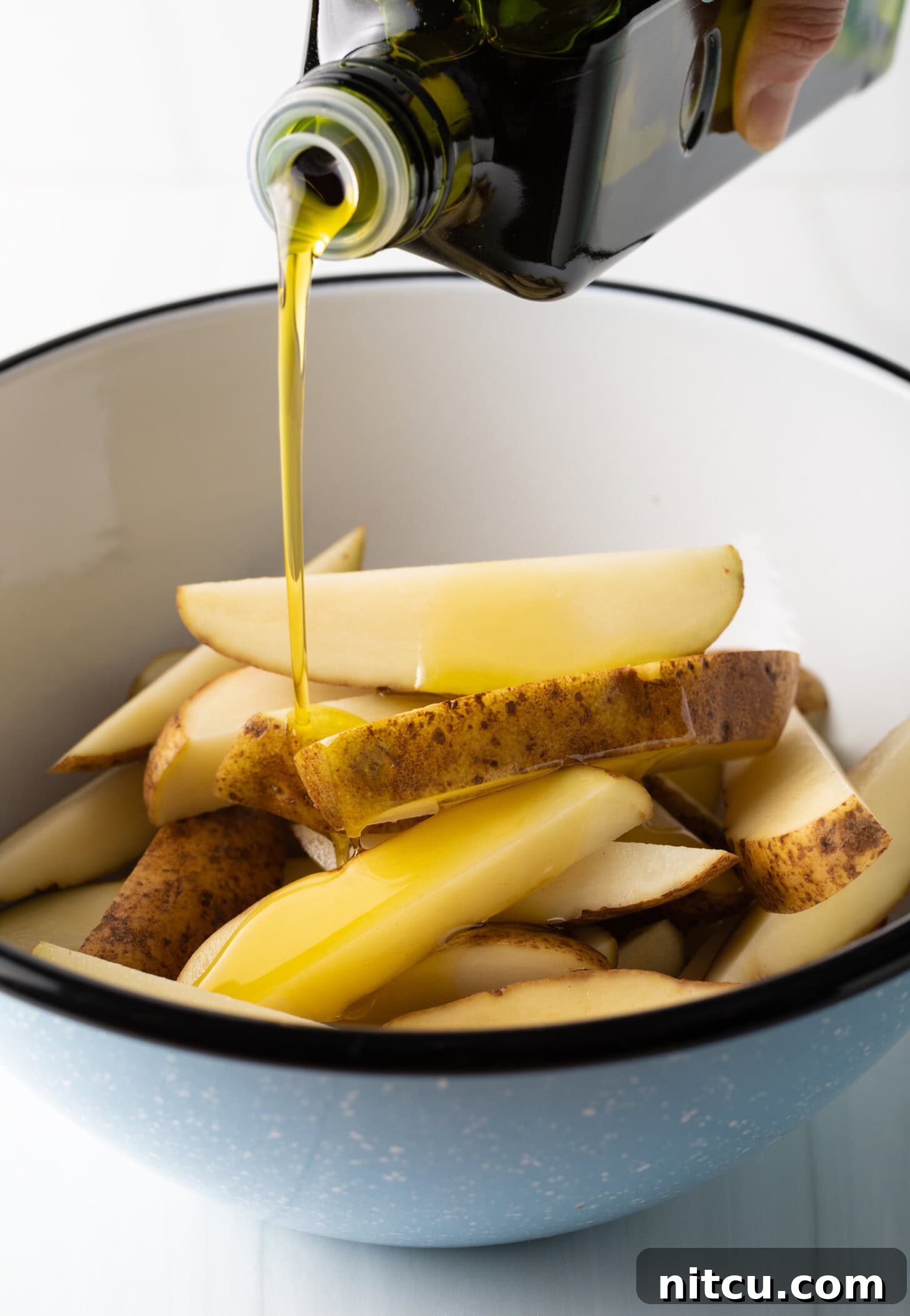 A hand is captured in the act of pouring olive oil from a bottle directly onto thick-cut potato wedges in a pristine white bowl, preparing them for the air fryer.