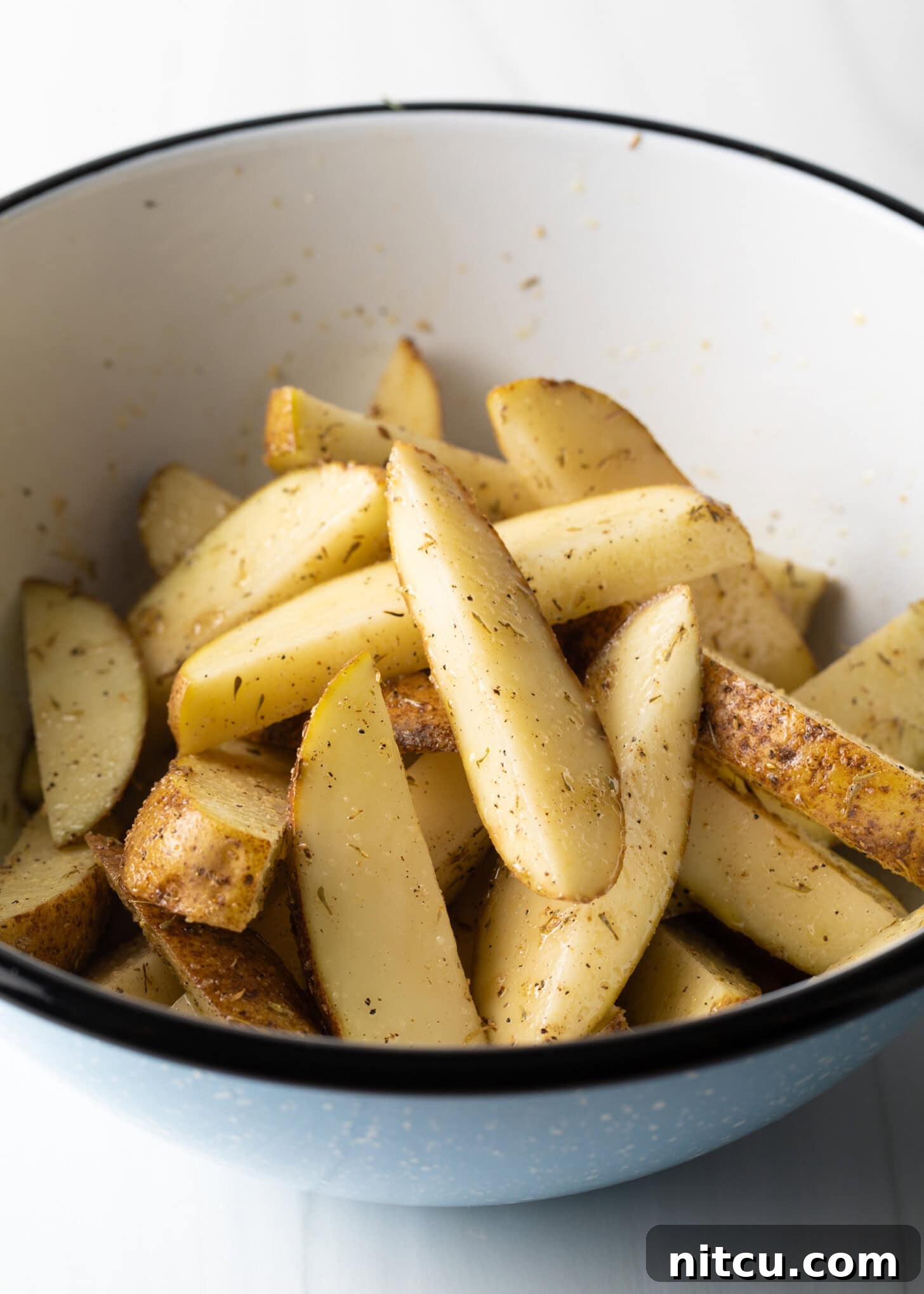 Thick-sliced raw potato wedges are visible in a white mixing bowl, perfectly tossed with olive oil and a vibrant blend of seasonings.