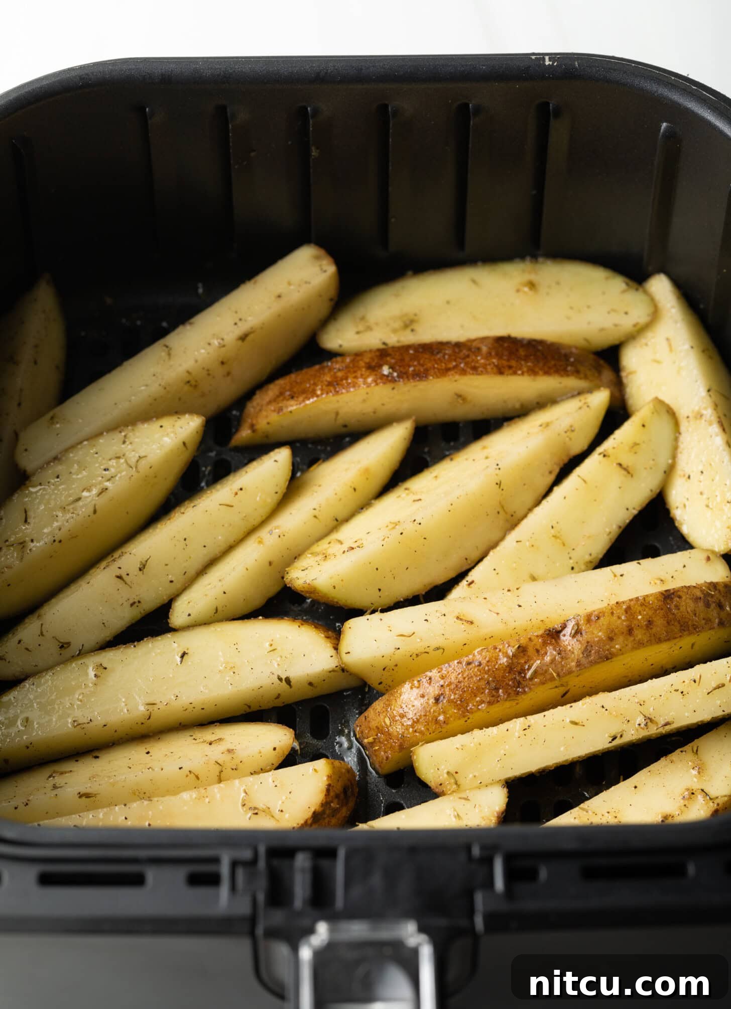 Raw potato wedges, cut into thick slices, are carefully spread in a single, even layer within the black basket of an air fryer, ready for cooking.